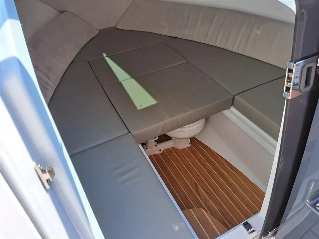 Interior of a boat cabin, featuring grey seating, wooden flooring, and a toilet, with sunlight casting a shadow.